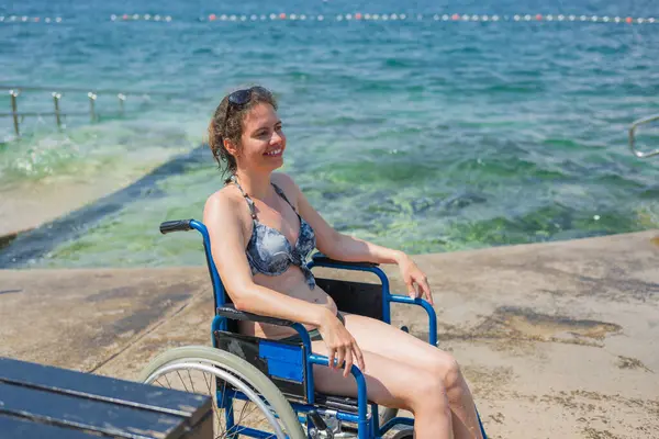 Bikinili gülümseyen bir kadın, deniz kenarında mavi tekerlekli sandalyesinde oturuyor, yaz tatilinde, bir deniz banyosundan sonra güneşin tadını çıkarıyor..