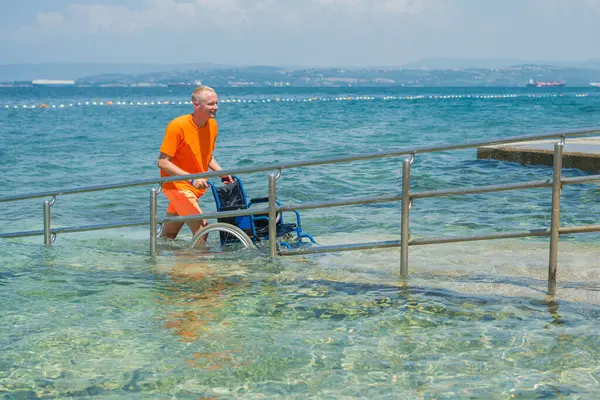Gülümseyen cankurtaran, arka planda güzel bir deniz manzarası olan bir yaz turistik bölgesinde ulaşılabilir bir sahil rampası boyunca boş bir su tekerlekli sandalyesini yuvarlıyor..