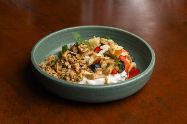 Yoğurt, taze meyve ve gevrek gevreğiyle dolu renkli bir brunch kasesi ahşap bir masada oturup besleyici ve iştah açıcı bir kahvaltı sunar..