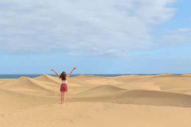 Altın kum tepelerinde çıplak ayakla duran bir insan, kutlamada kollarını kaldırmış, arkada okyanus parçalı bulutlu bir gökyüzünün altında görünür bir şekilde..