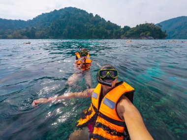 Parlak can yeleği giyen iki kişi berrak sularda şnorkelle yüzüyor. Çevreleri yemyeşil dağ manzaralarıyla çevriliyken canlı bir sualtı dünyasını keşfediyorlar..