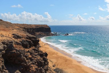 Parlak güneş ışığı, kayalık kayalıklarla çevrili kumlu sahili ve açık bir günde okyanusun yumuşak dalgalarını vurgular..