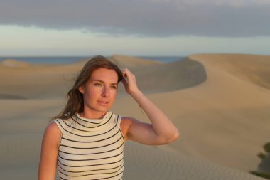 Bir kadın günbatımında kumlu bir çöl manzarasında düşünceli bir şekilde duruyor. Saçlarında hafif rüzgarlar ve etrafını saran kumdan tepeler var..