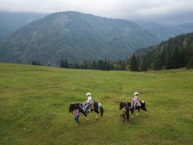 Yeşil bir çayırda at sürmekten hoşlanan bir çift, eğitmenlerin rehberliğinde, görkemli dağ manzaraları ve doğanın huzuru, havadan çekim..