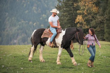 Kovboy şapkalı bir adam çimenli bir arazide siyah beyaz bir ata biniyor. Bir kadın yanında yürüyor, gün içinde atları dizginliyor..