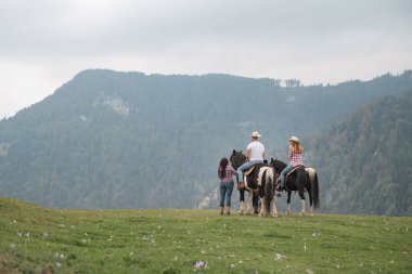 Avusturya Alpleri 'ndeki bir tarlada iki atlı üç kişi var. Bir kişi bulutlu bir günde arkasında dağlar olan bir biniciye yardım eder..