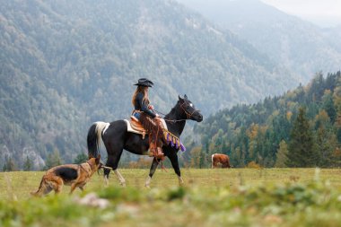 Kendine güvenen genç bir kadın, özgürlük ve macera ruhuna sahip bir kovboy kız muhteşem siyah bir ata biniyor, nefes kesici dağ arazisinde.