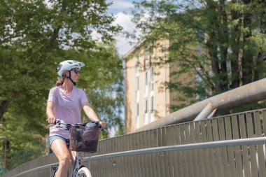 Parlak, güneşli bir günde, ağaçlar ve binalarla çevrili bir patika boyunca bisiklet süren kasklı bir kadın, yolculuğunun tadını çıkarıyor..