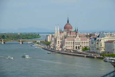 Budapeşte 'deki Tuna Nehri kıyısındaki etkileyici Macar Parlamentosu binası, açık mavi gökyüzü altında mimari güzelliğini sergiliyor. Tekneler su boyunca yavaşça süzülür..