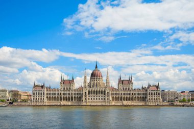 Budapeşte Parlamentosu binası Tuna Nehri boyunca parlak mavi bir gökyüzünün altında gururla duruyor. Ziyaretçiler, bölgenin çarpıcı mimarisi ve canlı atmosferinin keyfini çıkarıyorlar..