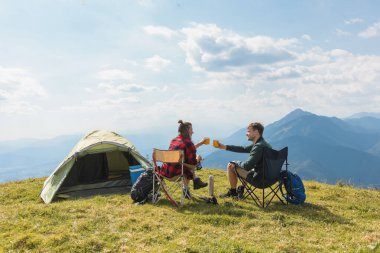 Çadırlarının yanındaki kamp sandalyelerinde dinlenen çift, manzaralı dağ manzarasının ve mavi gökyüzünün altında huzurlu bir atmosferin tadını çıkarıyorlar..