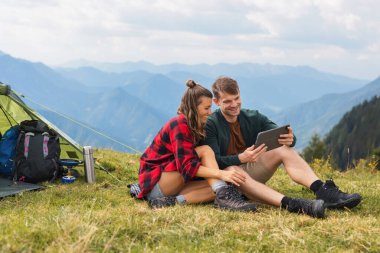Modern genç çift dağlarda kamp yapıyor, teknoloji ve doğayı harmanlıyor, dışarıda yemek pişirirken tablet üzerinde çalışıyor ve manzaranın tadını çıkarıyor..
