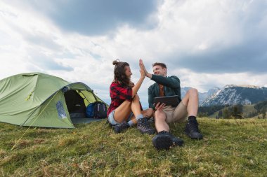 İki kampçı dağlarda neşeli bir anı paylaşıyor, bir çadırla çevrili çimlerin üzerinde otururken sohbet ediyorlar..