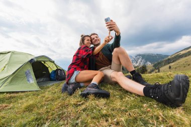Dağlarda kamp yapan bir çift, arka planda nefes kesici bir manzarayla selfie çekmek için akıllı telefon kullanıyor..