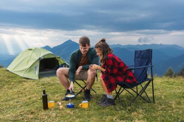 Genç bir çift kamp ocağında çırpılmış yumurta pişiriyor, manzaralı bir dağ kampında birlikte gülüyorlar, ve açık hava sabahının tadını çıkarıyorlar..