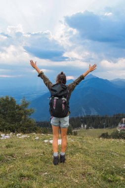 Sırt çantalı bir insan, kollarını bir dağın zirvesine kaldırmış bulutlu bir gökyüzünün altında nefes kesen manzaranın tadını çıkarıyor..