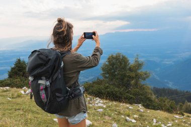 Sırt çantalı genç bir kadın eğlence gezisi sırasında akıllı telefonuyla güzel, sisli bir dağ manzarası çekiyor..