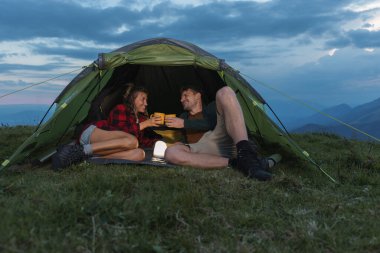 Bir çift, gün batımında manzaralı dağ manzarasıyla çevriliyken, sıcak içecekler ve gülümsemeler paylaşarak, bir dağdaki çadırda dinleniyor..