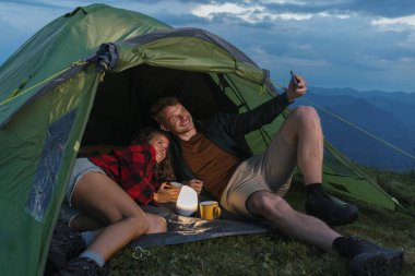 Bir çift çadırın içinde dinlenir, birlikte bir anı yakalarken, alacakaranlıkta çarpıcı dağ manzaralarıyla çevrilidir..