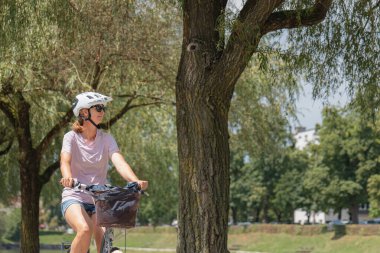 Genç bir kadın, güneşli bir günde, kaskı ve günlük kıyafetleriyle nehrin kenarındaki ağaçlık patika boyunca bisikletini sürüyor..