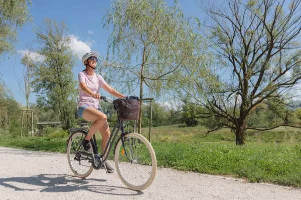Güneşli bir günde ağaçlarla çevrili çakıllı bir patika boyunca bisikletini süren bir kadın. Kask takıyor ve temiz havada gülümsüyor..