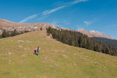 İki yürüyüşçü arka planda dağlarla çimenli tepelerde gezinir, maceralarının açık bir gününün tadını çıkarırlar..