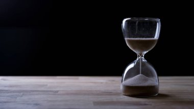 Kum saati, ahşap bir masada, koyu bir zemine karşı durur. Kum tepeden aşağı doğru akar..