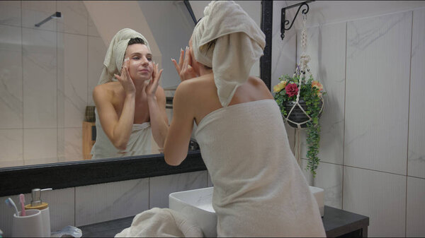 Young woman gazing into the bathroom mirror, applying skincare product to her face after her shower, with a towel on her head and wrapped around her body.
