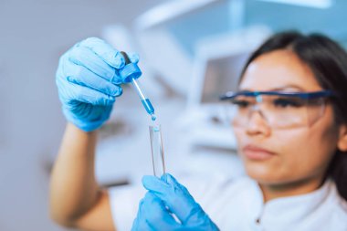 Koruyucu giysiler içinde kadın laboratuvar araştırmacısı, güvenlik gözlükleri ve mavi eldivenler dikkatlice sarı sıvıyı test tüpüne yerleştiriyor..