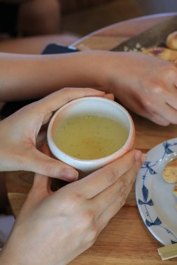 Japon restoranındaki ahşap masa arkasında bir fincan yeşil çay tutan kadın elleri. Küçük bir bardakta çay servisi.