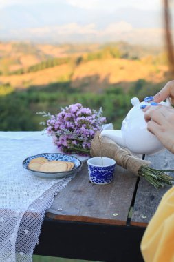 El yapımı çay beyaz çaydanlıktan bisküvi ve menekşe çiçekli ahşap masa manzaralı dağ manzaralı arka plan.