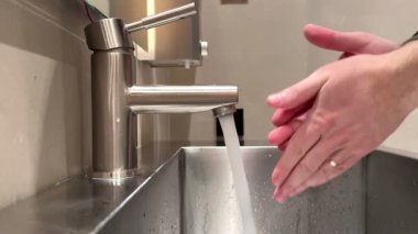Bir adam ellerini yıkamak için sıvı sabun kullanır ve sonra ellerini gümüş alüminyum lavaboda sıcak suda yıkar. Yüksek kalite 4k görüntü