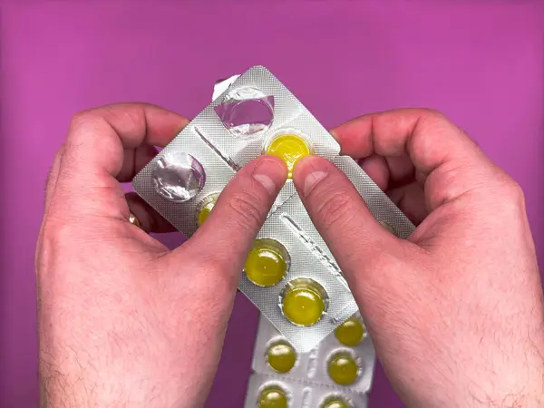 Erkek beyaz eller, parlak mor arka plandaki virüsü içmek ve tedavi etmek için gümüş bir paketten parlak sarı bir hap sıkarlar. Yüksek kalite fotoğraf