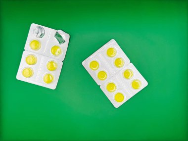 Gümüş ambalajlı iki paket parlak sarı yuvarlak tablet görüntünün ortasındaki parlak yeşil arka planda yatar. Bir paket açık üstten görüntüdür. Yüksek kalite fotoğraf