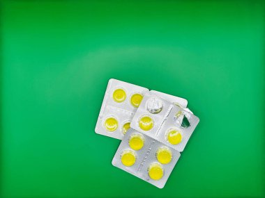 Gümüş ambalajlı parlak sarı yuvarlak tabletlerden yapılmış iki paket ilaç üst üste parlak yeşil bir zemin üzerinde uzanıyor, bir paket açık, üst görünüm. Yüksek kalite fotoğraf