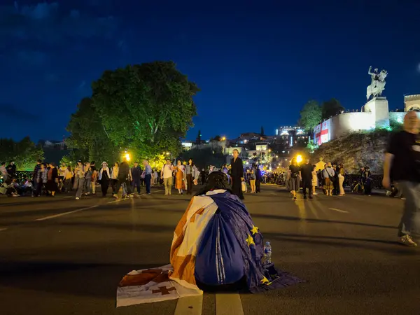 Tiflis, Gürcistan - 09 Mayıs 2025: Bir protestocu Gürcistan 'daki bir gece gösterisinin önünde arkasında Gürcistan ve Avrupa bayrağıyla yolda oturuyor. Yüksek kalite fotoğraf