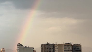 Bir şehrin üzerinde gökkuşağı görülür. Gökkuşağı uzun ve gökyüzü boyunca uzanıyor.