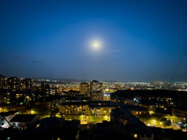 Şehir geceleri gökyüzünde parlayan bir dolunayla aydınlanıyor. Ay gökyüzünün ortasında konumlanmış, şehrin üzerine yumuşak bir parıltı yayıyor.