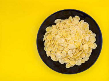 Bir kase sarı mısır gevreği. Gevrekler kasenin her tarafına saçılmış. Kase siyah renkli. Sarı zemin üzerinde mısır serisi. Kopya alanı, üst manzara, deri döşeme.