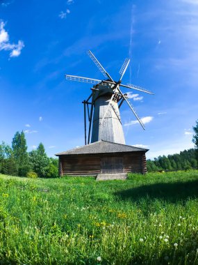 Bir yel değirmeni bir çimen tarlasının ortasındadır. Yel değirmeni ahşap bir binayla çevrili.