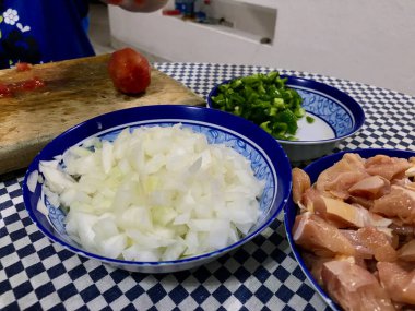 Kareli bir masa örtüsünde, içinde doğranmış tavuk, domates, biber, soğan ve sarımsak bulunan malzemeler pişiriyorum..