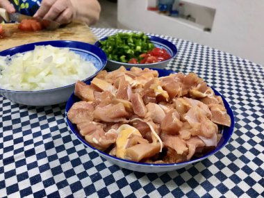 Kareli bir masa örtüsünde, içinde doğranmış tavuk, domates, biber, soğan ve sarımsak bulunan malzemeler pişiriyorum..