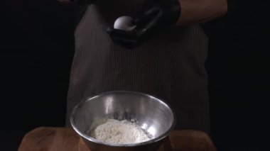 Erkek elleri una yumurta kırıyor, yakın plan. Şef hamur, ahşap masa ve siyah arka plan hazırlıyor. Pişirme evi kavramı. Yavaş çekim 