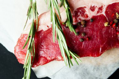 Raw organic beef meat with rosemary, seasonings, salt and red pepper  on black background, close-up. Preparing to cooking steak