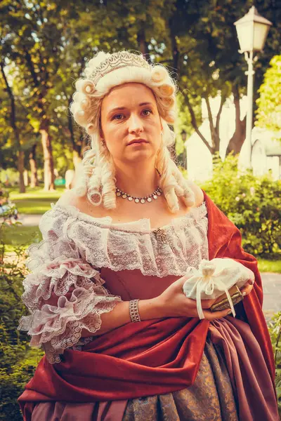 Portrait of blonde woman dressed in historical Baroque clothes with old fashion hairstyle, outdoors. Red color dress. Luxurious medieval dress