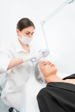 Kozmetik salonunun genç bayan müşterisi yüz maskesi takıyor. Yüzüne soyulan bir maske takma prosedürü. Cilt bakımı ve güzellik salonu veya klinik kavramları.