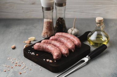 Kesme tahtasının üstünde ızgara sosis ızgara çatal, tuz ve biber, gri arka plan, kapat. Sağlıksız yiyecekler. Pişirme hazırlığı