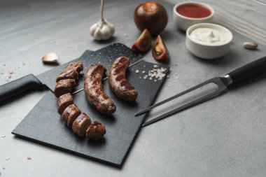 Kesme tahtasında ızgara çatal ve bıçakla ızgara sosis yemeye hazır. Gri arka plan, kapat. Sağlıksız yemek.