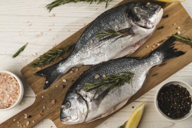 Limonlu taze dorado balığı, ahşap kesme tahtasındaki biberiye ve baharatlar, yakın çekim. Beyaz ahşap masa üzerinde malzemeli çiğ deniz ürünleri. Sağlıklı gıda konsepti