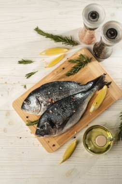 Limonlu taze dorado balığı, ahşap kesme tahtasındaki biberiye ve baharatlar, yakın çekim. Beyaz ahşap masa üzerinde malzemeli çiğ deniz ürünleri. Sağlıklı gıda konsepti